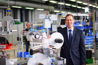 Un homme en costume se tient à côté d'une machine dans une usine, tandis qu'en arrière-plan, d'autres employés sont occupés.