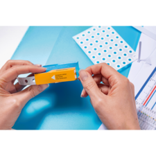 Eine Person hält eine Verpackung mit einem gelben Etikett in der Hand, während sich im Hintergrund ein Blatt mit Löchern und Diagramme befinden.