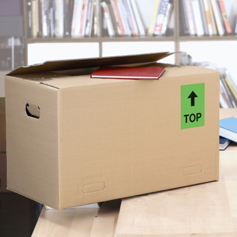 Kartonbox mit einem roten Heft auf dem Deckel, beschriftet mit einem grünen "TOP"-Schild, steht auf einem Tisch vor einem Bücherregal.