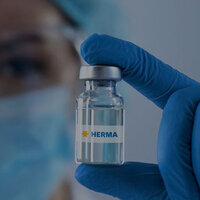Une personne portant des gants bleus tient une petite bouteille en verre avec l'inscription "HERMA".