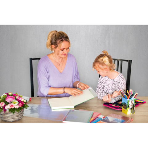 Eine Frau und ein Mädchen sitzen an einem Tisch und blättern in einem Buch. Auf dem Tisch liegen verschiedene Schreibmaterialien und Blumen.