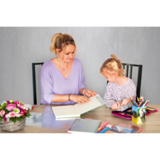 Eine Frau und ein Mädchen sitzen an einem Tisch und blättern in einem Buch. Auf dem Tisch liegen verschiedene Schreibmaterialien und Blumen.