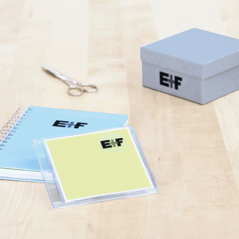 Ein blauer Notizblock, eine gelbe CD-Hülle mit dem Logo "E!F" und eine graue Geschenkbox auf einem Holztisch. Schere daneben.