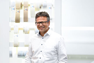 Un homme portant des lunettes et une chemise se tient souriant devant un fond lumineux et moderne composé de différents matériaux.