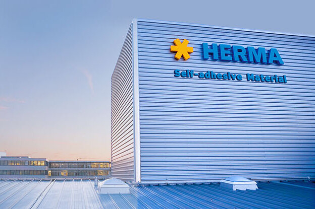 Le bâtiment avec l'inscription "HERMA" et un ciel bleu en arrière-plan. L'architecture est moderne et fonctionnelle.