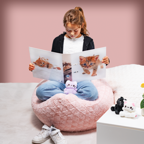 Un fille est assise dans un grand pouf moelleux et regarde un livre avec des images de chatons. Des peluches sont éparpillées autour d'elle.