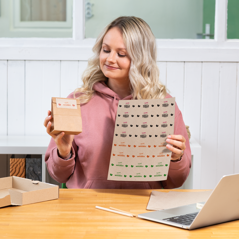 Eine Person mit blonden, lockigen Haaren hält eine kleine Verpackung und ein Blatt mit Aufklebern in einer hellen Umgebung.