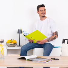 Ein junger Mann mit tätowierten Armen sitzt an einem Tisch, hält eine leuchtend grüne Mappe und schaut auf Dokumente.