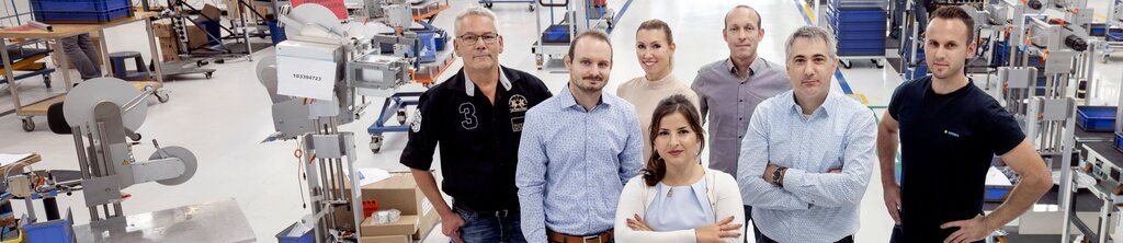 Une groupe de sept personnes se tient dans une salle de production, entouré de machines et de postes de travail. Elles regardent vers la caméra.