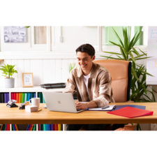 Ein junger Mann sitzt an einem Schreibtisch mit einem Laptop, umgeben von Schreibmaterialien und Pflanzen, und arbeitet konzentriert.