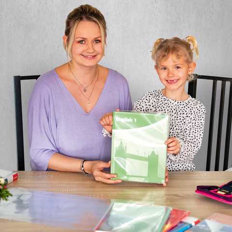 Eine Frau und ein Mädchen sitzen an einem Tisch, halten ein grünes Heft und lächeln. Auf dem Tisch liegen Schreibwaren und ein Blumenstrauß.