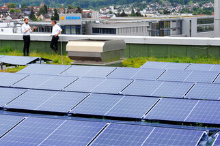 Deux personnes en costume se tiennent sur un toit équipé de panneaux solaires, avec des bâtiments et un paysage urbain visibles en arrière-plan.