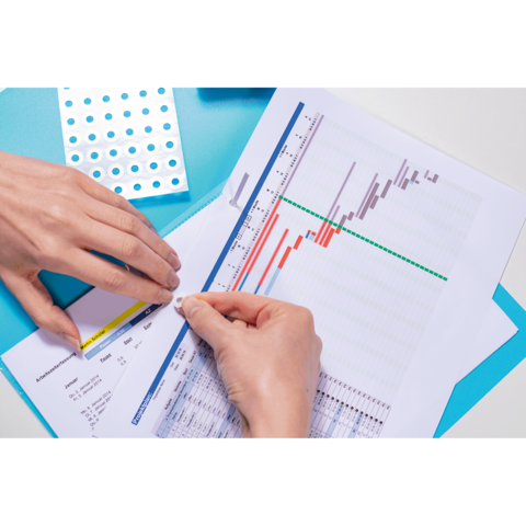 Hände, die ein Diagramm mit farbigen Balken und Linien auf einem Blatt Papier bearbeiten, während ein Aufkleber aufgebracht wird.