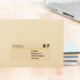 Ein gelbes Kuvert mit der Adresse D. Miller, Bergstr. 271, 70184 Stuttgart, Deutschland, liegt auf einem Tisch neben einem Laptop und Stapel von Papier.