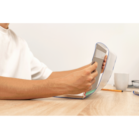 Eine Hand hält ein Smartphone, während die andere ein aufgeschlagenes Notizbuch auf einem Tisch hält. Ein Kaffeebecher ist im Hintergrund sichtbar.