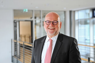 Un homme souriant, portant des lunettes et un costume, se tient dans un intérieur moderne. Il arbore une cravate rose et dégage une aura amicale.