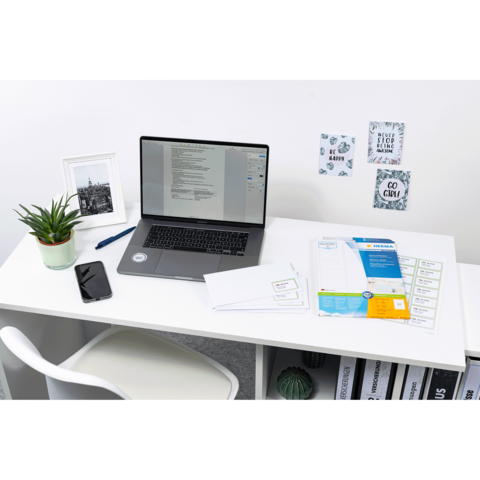 Ein Schreibtisch mit einem Laptop, einem Smartphone, einer Pflanze, Briefumschlägen und Dokumenten. An der Wand hängen dekorative Notizen.