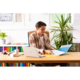 Ein junger Mann sitzt an einem Schreibtisch, arbeitet mit einem Laptop und einer Ordnermappe, umgeben von Pflanzen und bunten Akten.