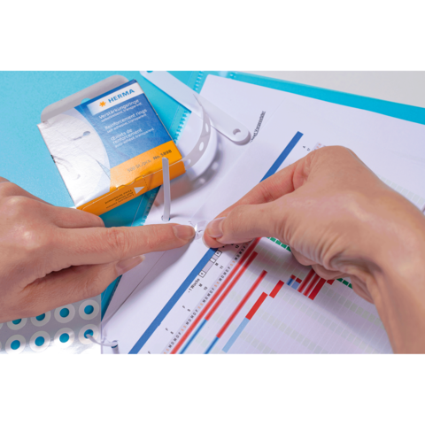 Eine Person befestigt eine transparente Kunststoffhülle an einem Dokument mit Diagrammen und Grafiken. Im Hintergrund ist eine Verpackung sichtbar.