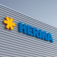 Des logos en forme d'étoile jaune et des lettres bleues forment le mot "HERMA" sur la façade d'un bâtiment moderne.