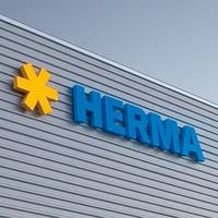 Des logos en forme d'étoile jaune et des lettres bleues forment le mot "HERMA" sur la façade d'un bâtiment moderne.