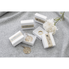 Schmuckstücke in weißen Geschenkboxen, darunter ein Baum-Anhänger, ein Ring und eine Blume auf einem grauen Untergrund.