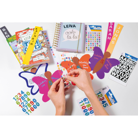 Hände, die bunte Aufkleber auf Schmetterlingsformen anbringen, umgeben von Schulmaterialien, Notizbüchern und Aufklebern mit Namen.