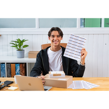 Ein junger Mann sitzt an einem Tisch, hält ein Blatt mit Aufklebern in der Hand und lächelt. Auf dem Tisch stehen ein Laptop und eine Kiste.