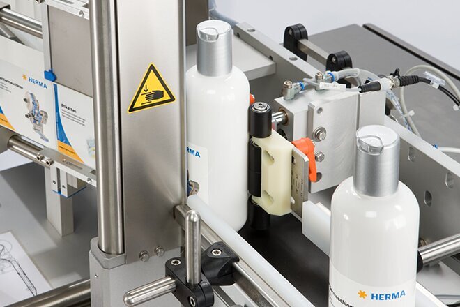 Une machine de remplissage avec des bouteilles blanches sur un tapis roulant, équipée de différents composants techniques et d'avertissements.