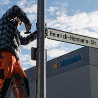 Un ouvrier fixe un panneau de rue portant l'inscription "Heinrich-Hermann-Str." à un poteau, tandis qu'un bâtiment est visible en arrière-plan.