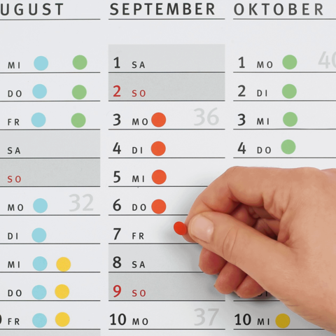 Ein Hand greift nach einem roten Punkt auf einem Kalender, der die Monate August, September und Oktober zeigt, mit markierten Tagen.