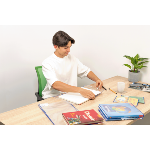 Ein junger Mann sitzt an einem Tisch, blättert in einem Notizbuch und hat Bücher sowie Schreibmaterialien vor sich. Eine Pflanze steht daneben.