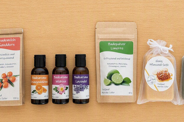 Étiquettes individuelles pour des produits cosmétiques en série : savons à base d'huile végétale dans des sachets transparents, Doypacks avec lait de bain et poudre de bain, ainsi que des bouteilles en verre brun avec arôme de bain, sur une surface en bois clair.