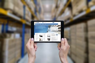 Texte : "Tablette en mains, il montre un site web avec des informations sur la gestion des stocks, en arrière-plan des étagères avec des cartons.