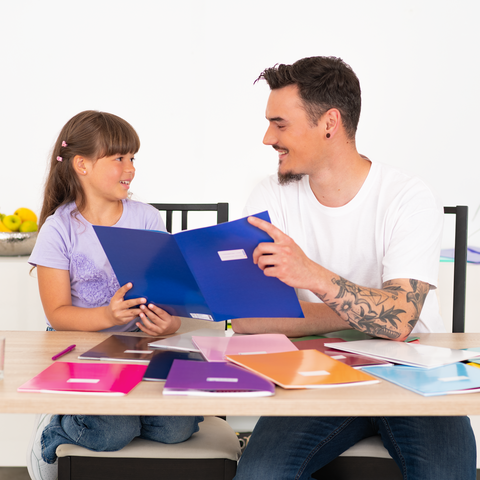 Ein Vater und seine Tochter sitzen an einem Tisch, umgeben von bunten Ordnern, während sie gemeinsam etwas besprechen.