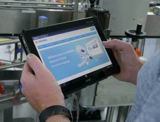 Une personne tient une tablette sur laquelle s'affiche le site web du portail de services HERMA, avec un message de bienvenue et un design de robot.