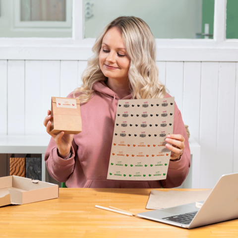 Eine Frau mit blonden, lockigen Haaren hält eine kleine Verpackung und ein Blatt mit Aufklebern in einem hellen Raum.