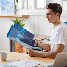 Ein junger Mann sitzt an einem Tisch und betrachtet eine Mappe mit Landschaftsbildern, während er eine Tasse Kaffee hält.