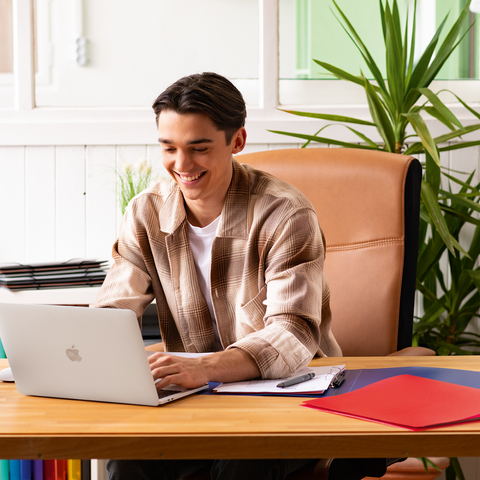 Ein junger Mann sitzt an einem Schreibtisch mit einem Laptop, umgeben von Büromaterialien und Pflanzen, während er arbeitet.