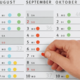 Ein Hand, die einen roten Punkt auf einem Kalenderblatt für September und Oktober anzeigt, mit markierten Daten für die Monate.