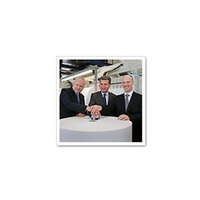 Trois hommes en costume se tiennent autour d'une table ronde et posent leurs mains sur un objet central.