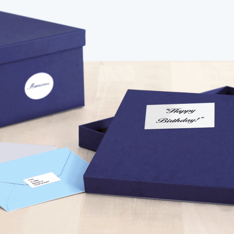 Eine geöffnete, dunkelblaue Geschenkbox mit einem "Happy Birthday!"-Aufkleber und zwei Umschlägen in hellblau und grau daneben.