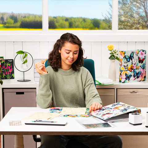 Une jeune femme est assise à une table, entourée de notes et d'un ordinateur portable, tandis qu'elle fait un geste de la main. En arrière-plan, des fenêtres offrent une vue sur un paysage verdoyant.
