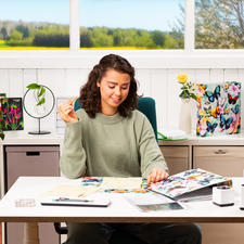 Une jeune femme est assise à une table, entourée de notes et d'un ordinateur portable, tandis qu'elle fait un geste de la main. En arrière-plan, des fenêtres offrent une vue sur un paysage verdoyant.