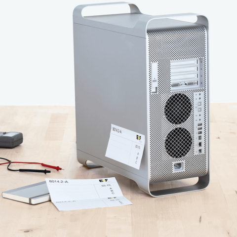 Silberner Computer-Tower mit zwei Lüftungsschlitzen, daneben ein Notizbuch und Messgeräte mit Kabeln auf einem Holztisch.