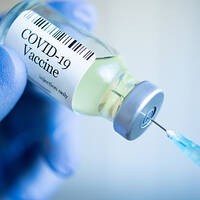 Une main gantée de bleu tient une bouteille de vaccin contre la COVID-19, tandis qu'une seringue est prête à prélever le liquide.