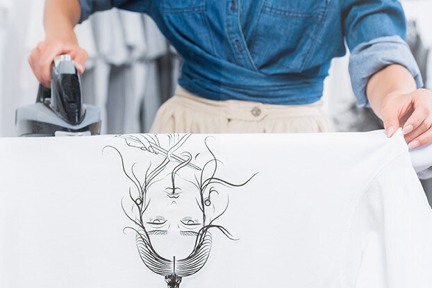 Une personne repasse une chemise blanche à motif graphique tout en se tenant sur une planche à repasser.