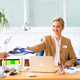 Une femme dans un bureau tend un document à un homme, tandis qu'elle est assise à un bureau avec un ordinateur portable et du matériel de bureau.
