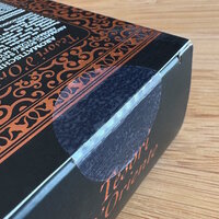 Solutions Cosmétiques : Emballage noir avec des motifs décoratifs orange et du texte. Le rabat de l'emballage est scellé avec une étiquette de fermeture transparente.