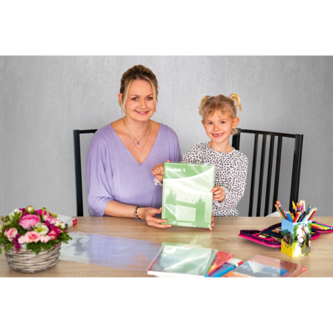 Eine Frau und ein Mädchen sitzen an einem Tisch und halten ein grünes Heft. Auf dem Tisch liegen Schreibmaterialien und Blumen.